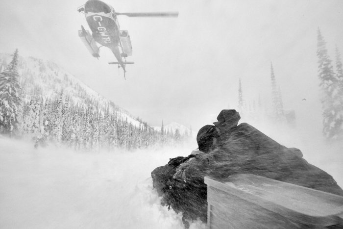 A skier huddles down against the surface of the snow and braces himself as a helicopter drops him off on peak during heliskiing.