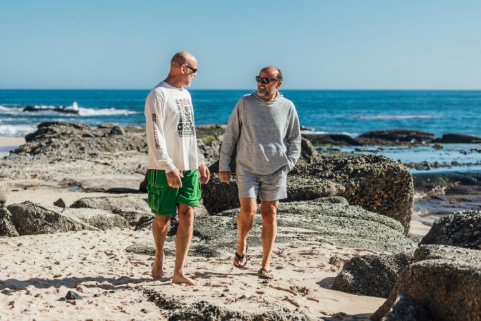 Two older gay men in shorts and sunglasses walk along an ocean beach in the sun.