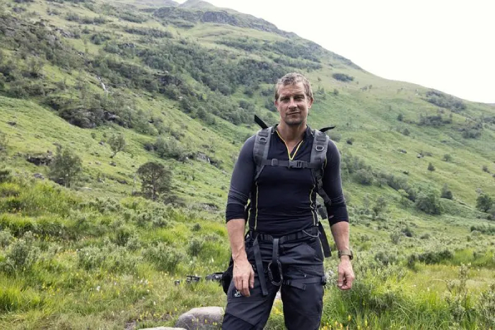 Bear Grylls wearing adventure gear stands in front of a green hillside.