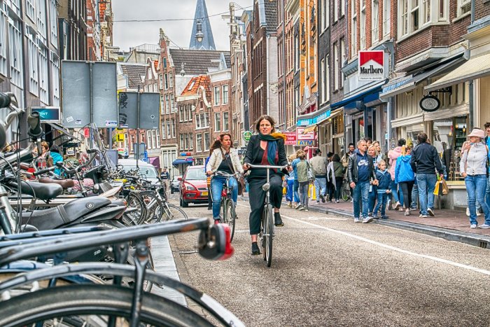 Bicyclists ride along a busy alleyway with parked bicycles and pedestrians shopping.