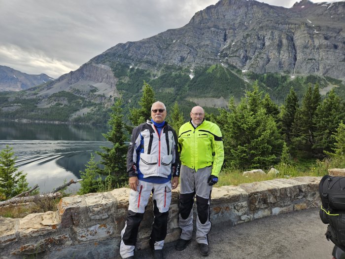 Two male motorcycle riders pose for a photo in front of a scenic mountain scene.