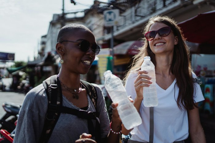 A young, black woman and a young, white woman drink bottled water in a city while traveling.