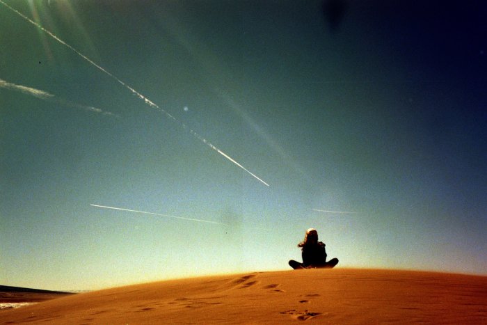 A woman sits cross-legged in the desert with contrails from planes above her.