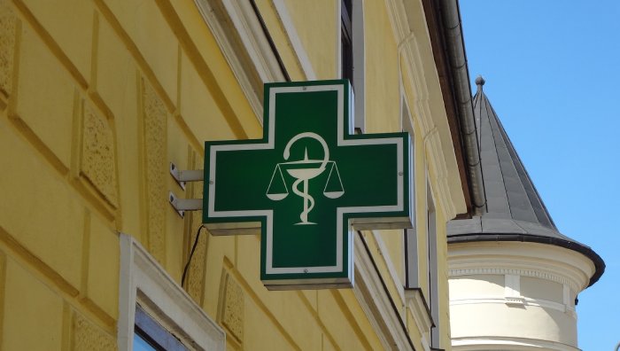 The green cross of a European pharmacy, mounted on the outside wall of a yellow building.