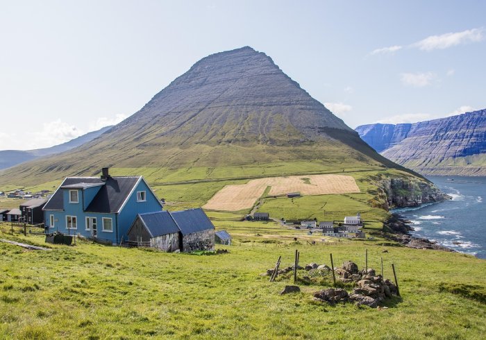 A blue house built on a grassy hillside overlooking the ocean with mountains and cliffs in the background.