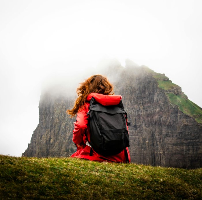 A woman with red hair and a red jacket and black backpack sits with her back to the camera on a grassy knoll and looks up at a tall, rocky, seaside mountain.