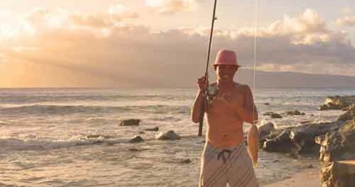 A shirtless fisherman in a bucket hat proudly smiles and shows off his fish.