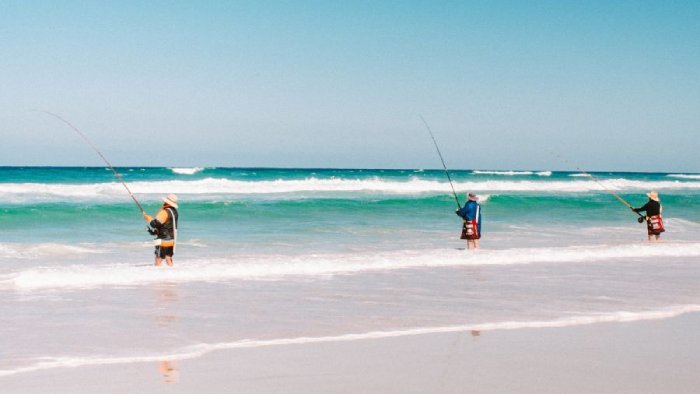 Three people stand in shallow ocean water near a beach to surfcast.