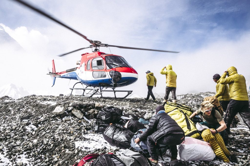 A rescue helicopter on a rocky landing pad with mountain rescue workers wearing yellow jackets loading equipment.