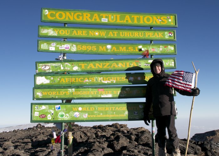A woman poses with an American flag in front of an enormous summit sign on top of a mountain in Africa.