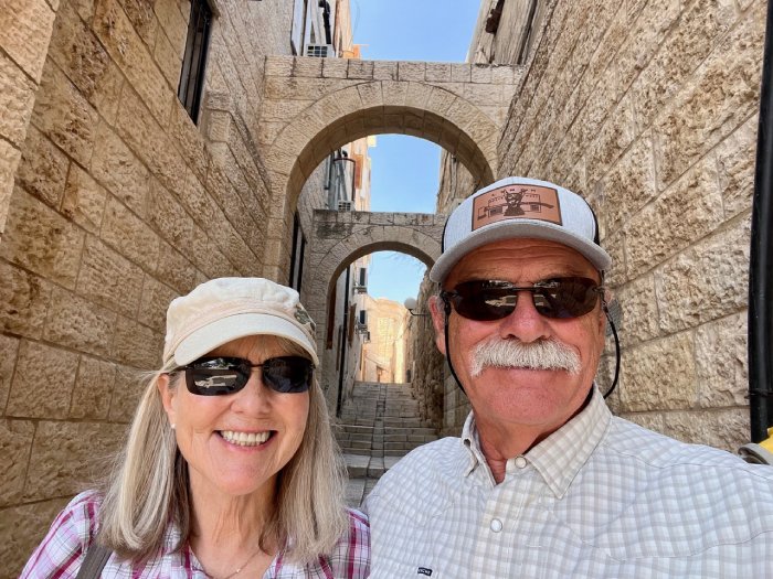 A middle-aged white couple wearing sunglasses and hats poses for a photo beneath arches in an Israeli city.