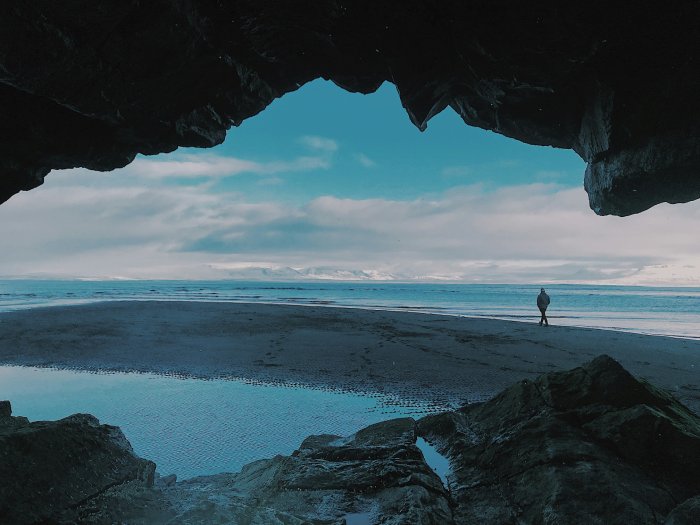 A man walks alone on a cold beach in Iceland with snow-capped mountains in the background.