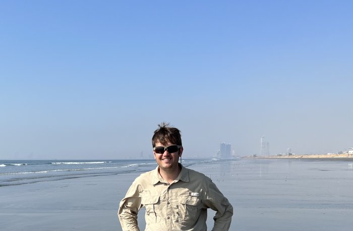 A man wearing sunglasses stands on a massive beach with skyscrapers in the background.