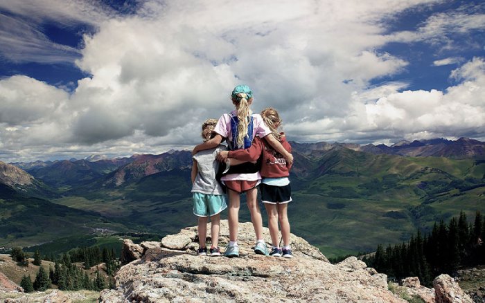 Three young sisters stand on top of a rocky point overlooking green mountains under a cloudy blue sky.