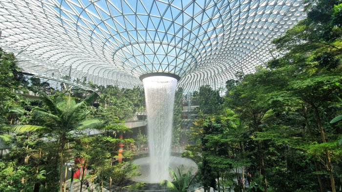 A man-made vortex waterfall flows from the glass ceiling into a lush room in an airport.