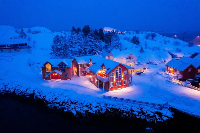 A cozy red house with many interior lights on illuminates its wintry surroundings on a snowy property in Lofoten, Norway at dusk.