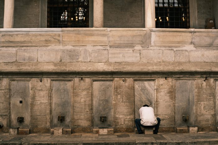 A man squats at a public water fountain in Turkey to fill a water bottle.