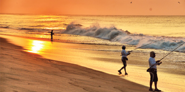 Three people surfcast on a Mexican beach under a setting sun.