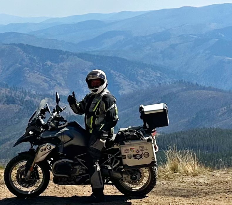 A motorcycle rider on his touring motorcycle poses for a photo in front of mountains.