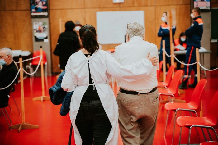 A nurse in protective clothing walks an elderly man in a room with health security guards and a red floor and chairs to an area to get a COVID shot.