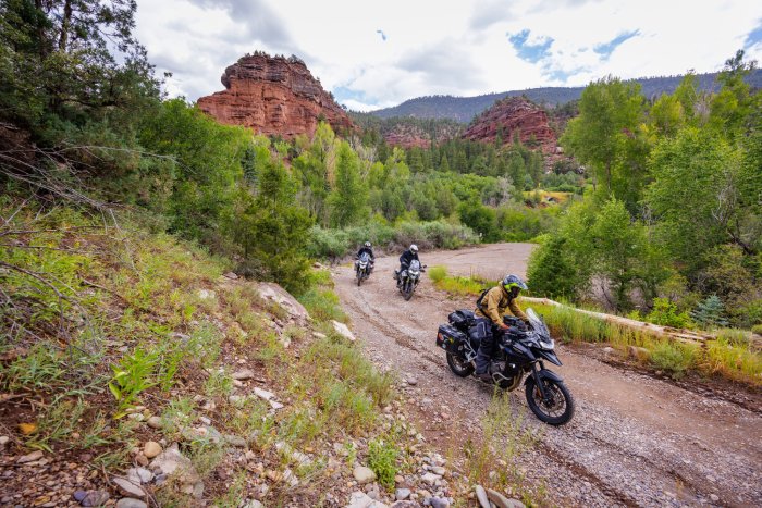 Three motorcyclists ride in the high desert, off-road among green plants and trees and red plateaus. 