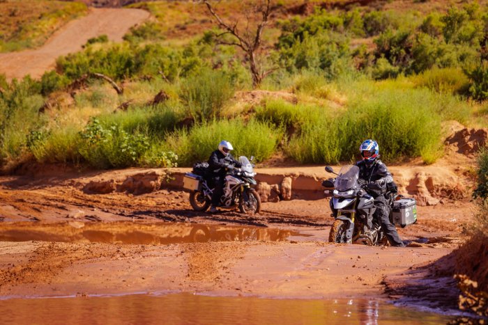 Two motorcyclists ride through deep, red mud in an attempt to cross a canyon river.
