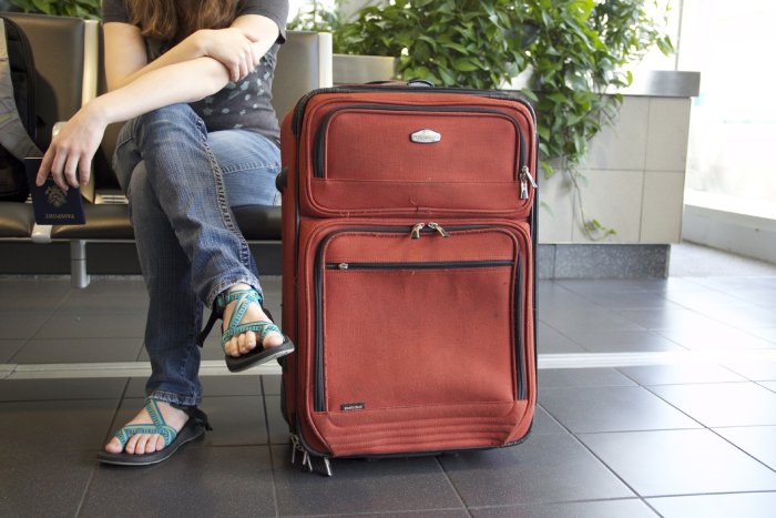 A well-used, terracotta-colored carry-on suitcase on the ground next to its passenger who is holding a passport while she waits for a plane.