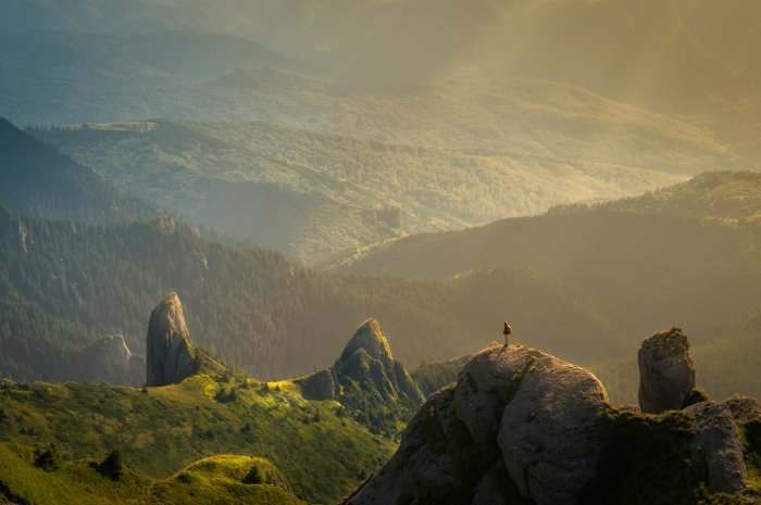 A person stands on top of a rocky overlook in the mountains looking out at the valley below.