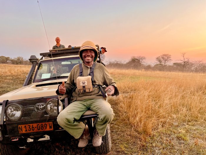 A smiling guide guide sits on the front of a safari jeep with smiling passengers in an African prairie.