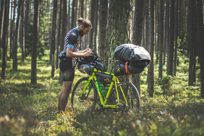 A bearded man in cycling clothes loads his bike in a forest during a bikepacking trip.