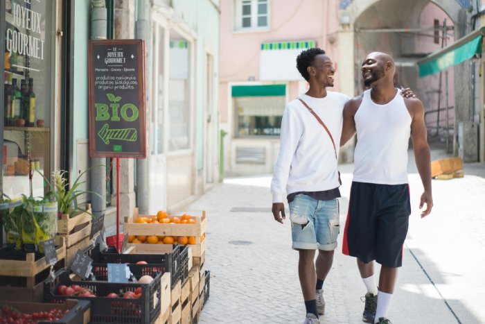 Two smiling, gay black men walk with arms around each other through a street market.