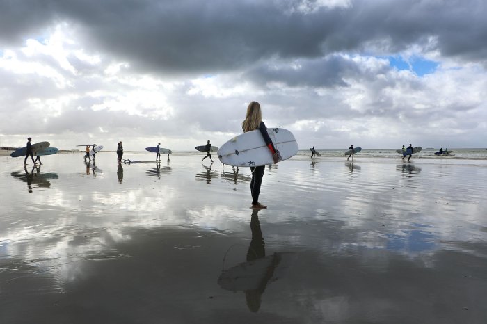 A surfing woman looks on as many other surfers walk out to the break during low tide on a mostly cloudy day.