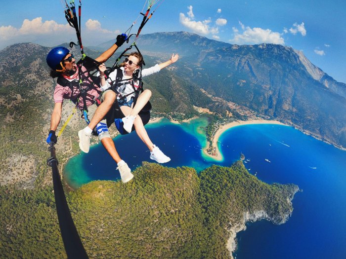 Tandem paragliders take a funny selfie high above a tropical island with mountains and clear blue water.