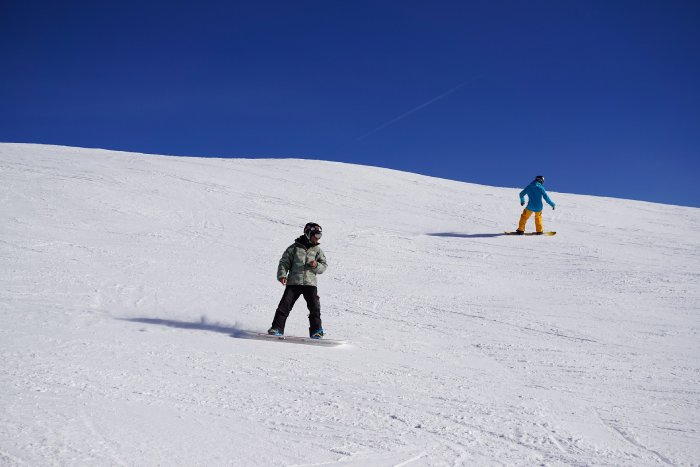 Two snowboarders ride down a snowy trail under a blue sky.