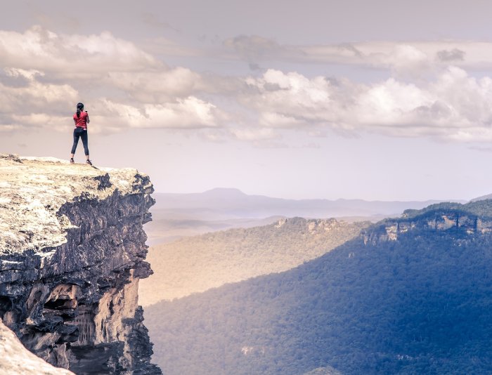 A young woman stands near the edge of a cliff overlooking tree-covered mountains.