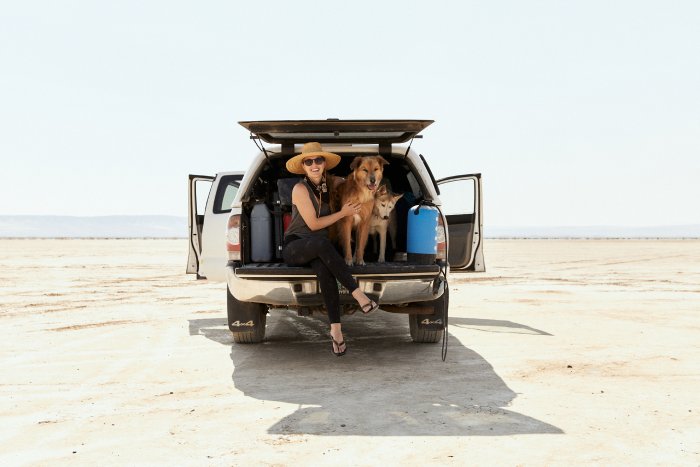 A woman sits on the tailgate of her truck with two dogs in the desert.