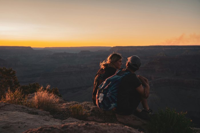 A woman and a man sit on top of a canyon at sunset.