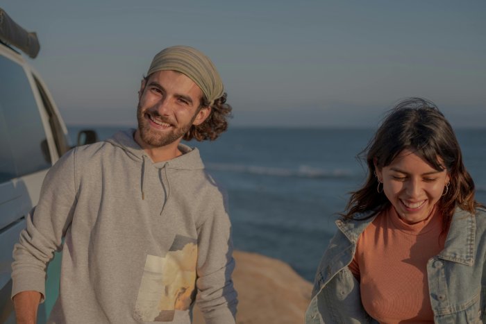 A young white man and a young white woman laugh near an SUV by the beach.