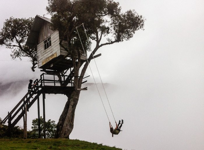 A woman swings from a tree swing with a treehouse above her in the clouds.