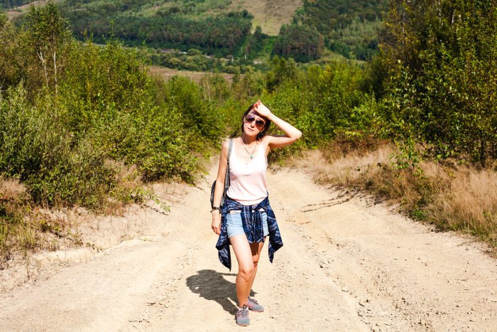 A tired, overheating young woman tourist hiking uphill in the hills on a hot, sunny day.