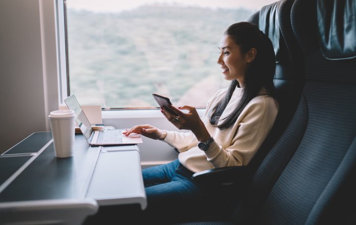 A young female business traveler on a train talks on her phone and works on her laptop.