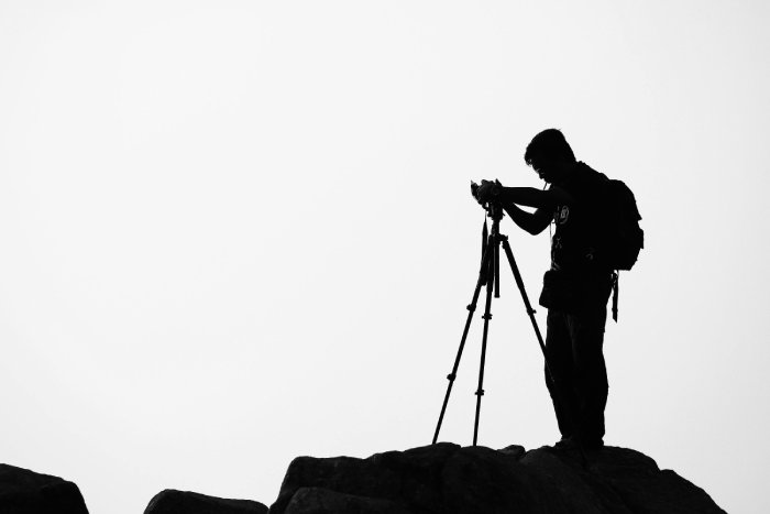 A silhouette of a man with a camera on a tripod standing on top of a rock.