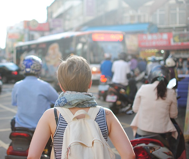 A college student wearing a backpack walks among the motorized scooters on a crowded street in Asia.