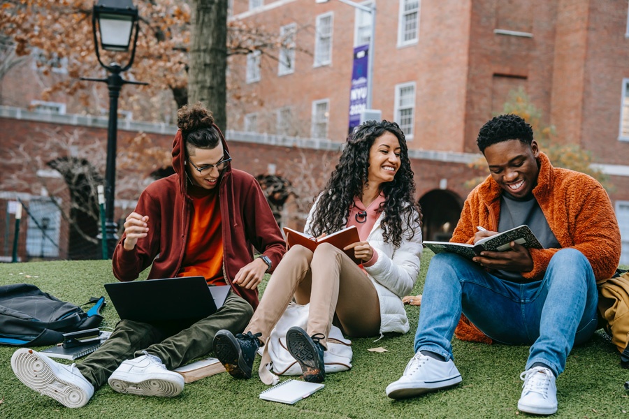 Three laughing college students sit and study outside on campus,