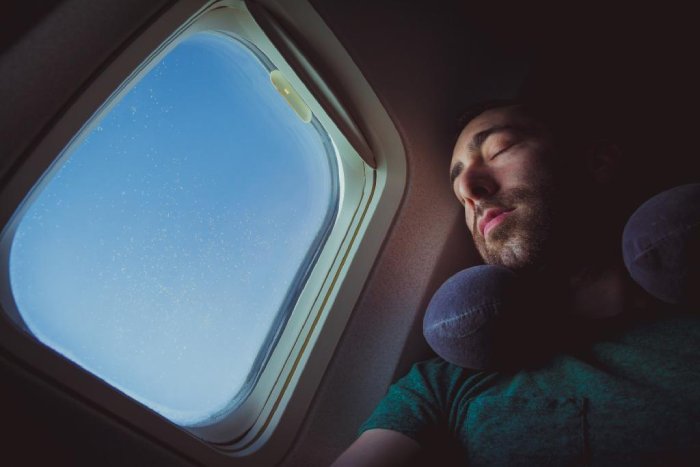 A man with a neck pillow rests his head with eyes closed against his window on a plane.