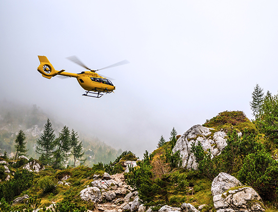 A yellow rescue helicopter approaches a landing zone atop a rugged mountain in cloudy conditions.