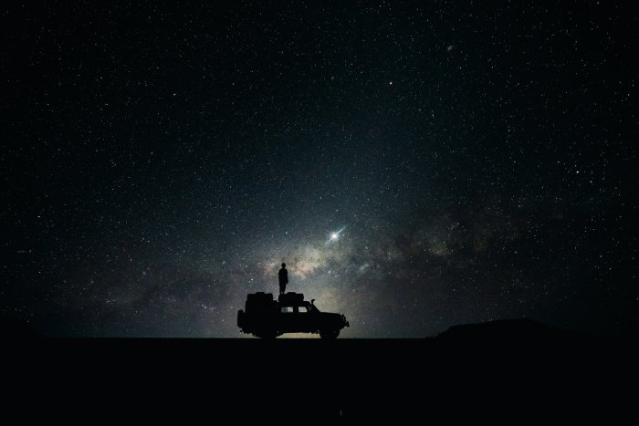 A man stands on the roof of his SUV in the dark looking up at the stars.