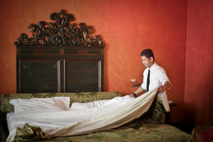 A man makes a large bed in a hotel room with red walls and a tall backboard.