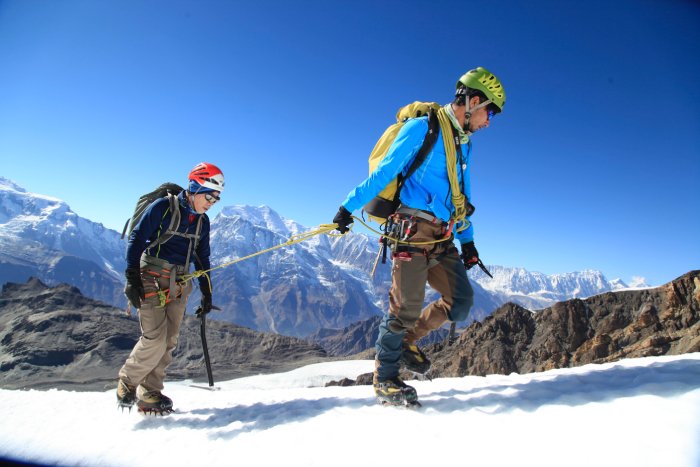 Two mountaineers roped together climb up a high-altitude slope under sunny skies.
