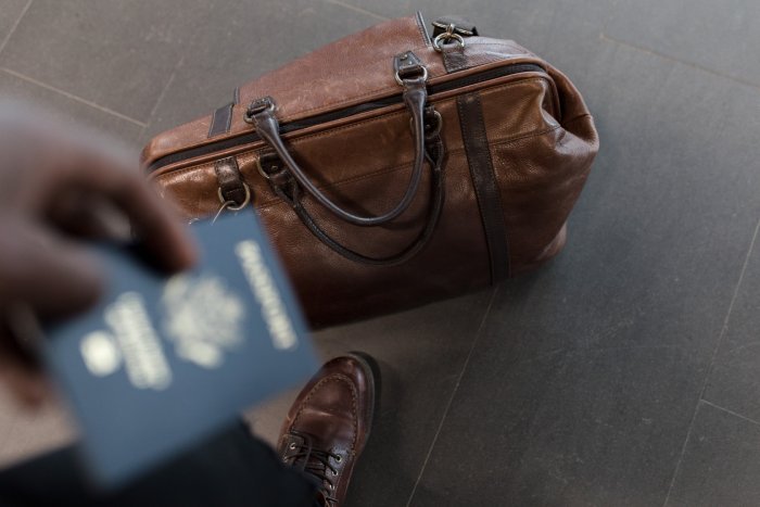 A person holds a passport with a brown travel bag on the ground next to him.